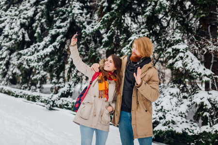 Young couple in love rest in park winter. A man hugs girlfriend, woman holds mobile phone in her hand and takes photo of herself. People use smartphone for selfie photos the winter in the park.の写真素材