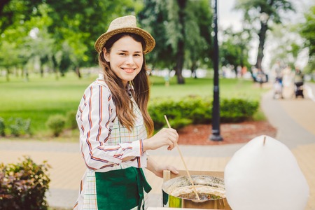photo theme small business cooking sweets. A young caucasian woman with an apron trader in the hat the owner of the outlet makes a candy floss, a fairy floss or a cotton candy in the summer park.の写真素材