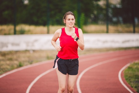 Beautiful young athlete Caucasian woman with big breasts in red T-shirt and short shorts running jog, running in a stadium with a red rubber coating. Hairstyle of ponytailの写真素材