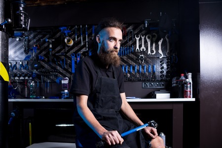 Portrait of small business owner of young man with beard. Guy bicycle mechanic workshop worker sitting with tool in his hand in a working black clothes in an apron in the background of a bicycle shop.の写真素材