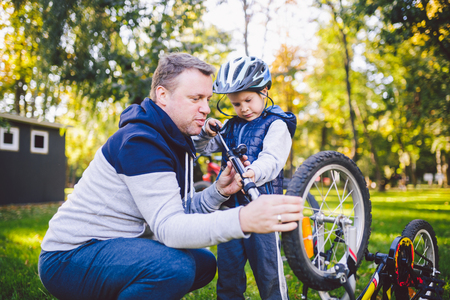 Father's day Caucasian dad and 5 year old son in the backyard near the house on the green grass on the lawn repairing a bicycle, pumping a bicycle wheel. Dad teaches how to repair a child's bike.の写真素材