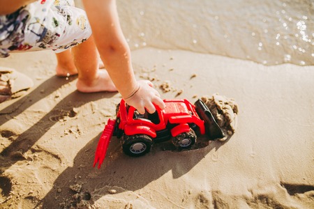 Caucasian child boy plays toy red tractor, excavator on sandy beach by the river in shorts at sunset...