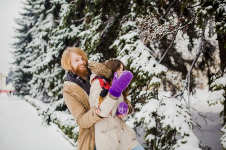 Young Caucasian boy with a beard and a girl have a date outdoors in the winter park against the background of a snowy conifer tree play snowballs, throw snow, hang themselves and play winter games.の写真素材