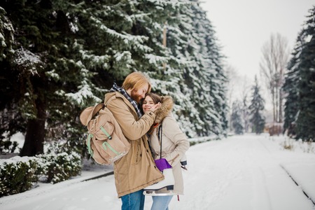Date young couple in winter. Young Caucasian man with beard and long hair holding in hands the hands of girl with knitted gloves and warming and kissing them in snow park in winter is Valentines Day.の写真素材