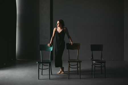 The social theme female loneliness pain suffering. Abastration male violence family. A young beautiful Caucasian woman in black clothes sitting on three chairs in large dark room. Srach and despair.の写真素材