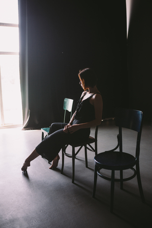 The social theme female loneliness pain suffering. Abastration male violence family. A young beautiful Caucasian woman in black clothes sitting on three chairs in large dark room. Srach and despair.の写真素材