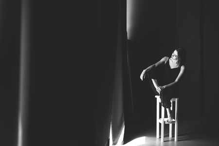 The social theme female loneliness pain suffering. Abastration male violence family. A young beautiful Caucasian woman in black clothes sitting on three chairs in large dark room. Srach and despair.の写真素材