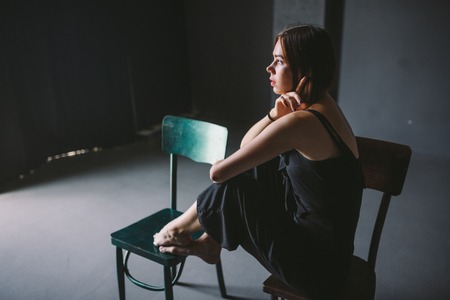 The social theme female loneliness pain suffering. Abastration male violence family. A young beautiful Caucasian woman in black clothes sitting on three chairs in large dark room. Srach and despair.の写真素材