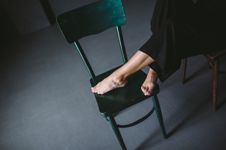 The social theme female loneliness pain suffering. Abastration male violence family. A young beautiful Caucasian woman in black clothes sitting on three chairs in large dark room. Srach and despair.の写真素材