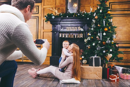 Theme mobile photography, amateur photo video phone. Hands Caucasian man holds uses smartphone makes photo mother and son home near fireplace and Christmas tree winter Christmas time New Year decor.の写真素材