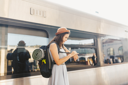 Theme transportation and travel. young Caucasian woman with toothy smile standing at station train background with backpack waving checks time on wrist hand wrist watch in dress and hat in summer.の写真素材