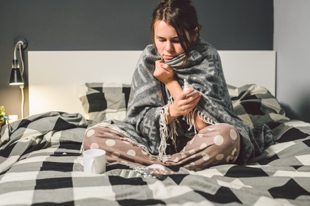 The theme is seasonal cold, runny nose flu virus infection. Young Caucasian woman at home bedroom bed sitting wrapping blanket, chill heat high temperature. Cup drink pills blister season flu.の写真素材