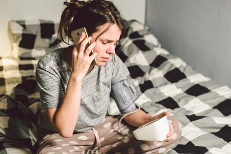 The topic high blood pressure hypertension disease. Beautiful young woman uses automatic tonometer to check pressure at home in the bedroom on the bed. A woman asks for help, call an ambulance 911.の写真素材