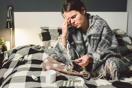 Subject disease and treatment. young Caucasian woman sits home bedroom bed wrapped in body blanket with high fever, fever holds strong headache behind head, uses drop medicine in nose for sinusitis.の写真素材