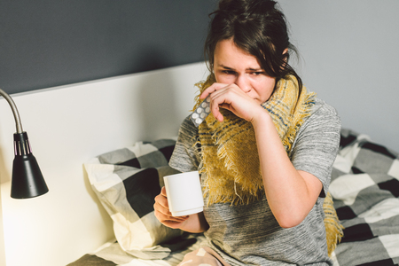 Young beautiful Caucasian woman has a cold, flu with high fever and heat at home in bed. The girl takes headache pills with a glass of water, a bitter pill is crooked.の写真素材