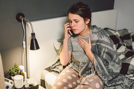 respiratory diseases and home remedies. young woman sick with cold sitting bedroom on bed holding throat sadness emotion. Sore throat man takes pills. Call phone, ask for help doctor. Call ambulance.の写真素材