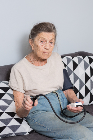 The topic is very old person and health problems. A senior Caucasian woman, 90 years old, with wrinkles and gray hair, sits home on sofa and uses a blood pressure monitor to measure blood pressure.の写真素材