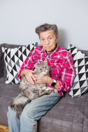 very old senior Caucasian grandmother with gray hair and deep wrinkles is sitting at home on the couch in jeans and a red checkered shirt with a gray fluffy shaggy cat in her arms. Pensioner and pet.の写真素材