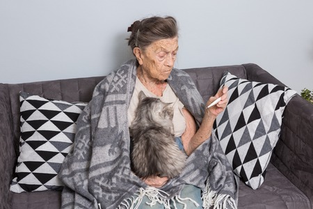 A very old senior Caucasian grandmother with gray hair and deep wrinkles is sitting at home on a sofa with a pet cat. Illness is cold and fever. Pensioner holds a thermometer in his hands.の写真素材