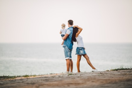Family holiday in nature. Three, Mom, Dad, daughter one year standing with backs on cliff overlooking sea. man holding hild in arms,woman hugging husband.Fashionable people dressed in stylish clothes.の写真素材
