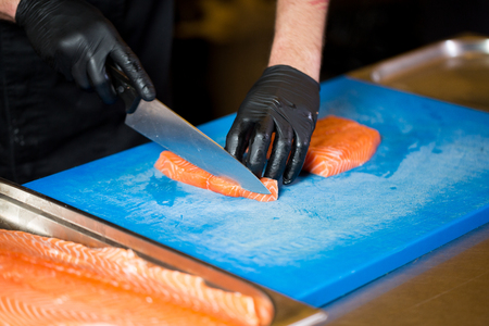 Theme cooking is a profession of cooking. Close-up of a Caucasian man's hand in a restaurant kitchen preparing red fish fillets salmon meat in black latex gloves uniform.の写真素材
