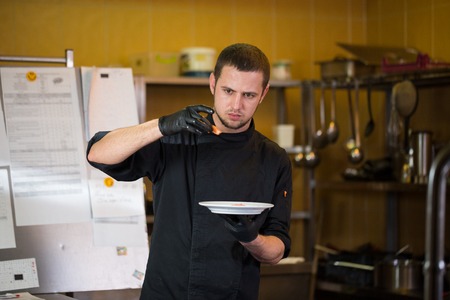 Theme cooking is profession of cooking. Portrait of Caucasian man in restaurant kitchen preparing red fish fillets salmon meat in black gloves uniform. Chef holds plate with blue and checks, tastes.の写真素材