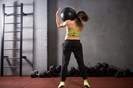 Theme sport and health. A strong muscular Caucasian woman in the gym trains strength and endurance. Equipping a large printed black ball raises and throws over her shoulder. Girl athlete crossfit.の写真素材