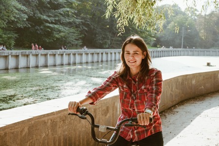 Subject ecological bicycle transport. Young caucasian woman on a dirt road in a park near a lake renting an orange-colored bike posing for a rest in the fall in sunny weather.の写真素材