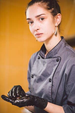 Subject profession and cooking pastry. young Caucasian woman with tattoo of pastry chef in kitchen of restaurant preparing round chocolate candies handmade truffle in black gloves and uniform.の写真素材