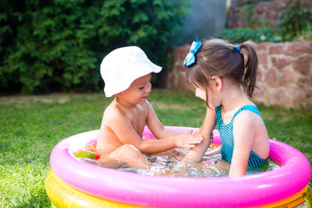 Theme is a children's summer vacation. Two Caucasian children, brother and sister, sit in a perched round pool with water in the yard of the green grass in a bathing suit and joy happiness smile.の写真素材