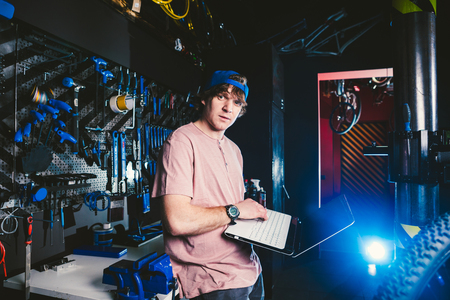 Theme business profession manager and technology. Young stylish Caucasian male owner bicycle shop uses a laptop computer near the stand with the tools for repairing and servicing the bicycle.の写真素材