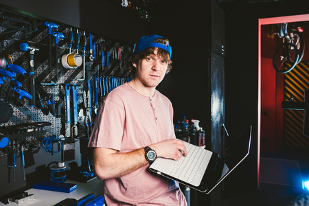 Theme business profession manager and technology. Young stylish Caucasian male owner bicycle shop uses a laptop computer near the stand with the tools for repairing and servicing the bicycle.の写真素材
