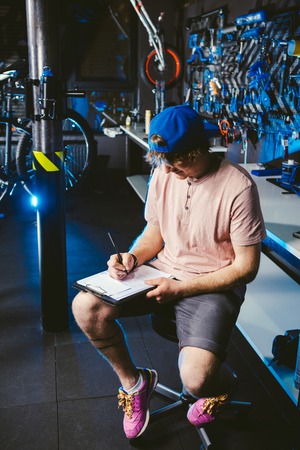 Young handsome stylish man in a cap snapback and with a tattoo small business owner selling a bicycle, the workshop sits on the background of the store in the hands of a notebook to write notes.の写真素材