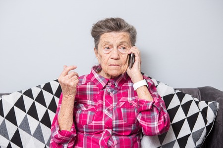 An old elderly woman grandmother with gray hair sits at home on the couch using the hand phone, a telephone conversation to hear the bad news. Emotion fear scare.の写真素材