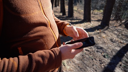 theme tourism and technology. Young caucasian man with beard and backpack. Hiking tourist in pine forest uses technology, hand holding mobile phone to touch the screen. Gps application orientation.の写真素材