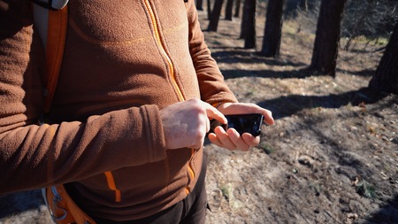 theme tourism and technology. Young caucasian man with beard and backpack. Hiking tourist in pine forest uses technology, hand holding mobile phone to touch the screen. Gps application orientation.の写真素材
