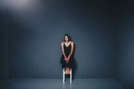 social theme of female loneliness is pain suffering. Abastration of male domestic violence. Young beautiful Caucasian woman in black clothes, strange pose, white chair big dark room. Fear and despair.の写真素材