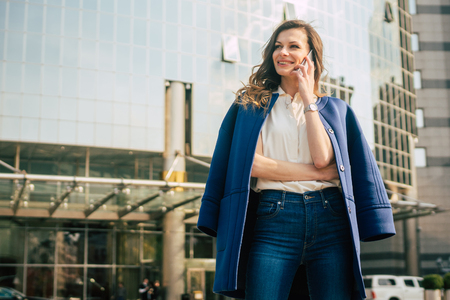 Caucasian business woman speaking by phone. Waist up portrait of a successful European business woman woman, talking on the phone, standing on glass background, modern office building. Sunny weather.の写真素材