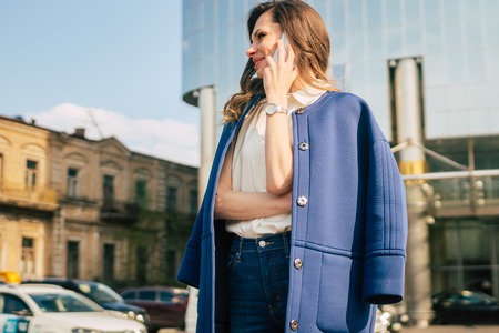 Caucasian business woman speaking by phone. Waist up portrait of a successful European business woman woman, talking on the phone, standing on glass background, modern office building. Sunny weather.の写真素材