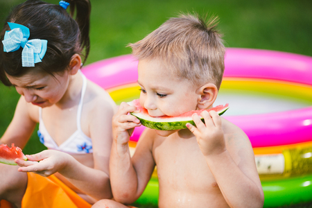 Funny little kids brother and sister eating watermelon on green grass near inflatable pool in yard at home.の写真素材
