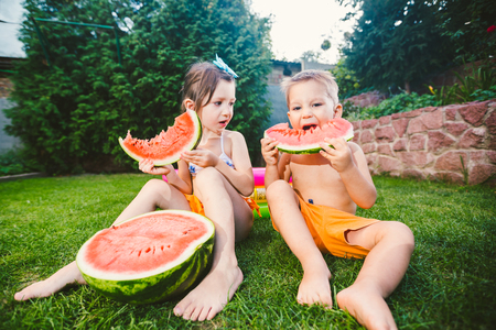 Funny little kids brother and sister eating watermelon on green grass near inflatable pool in yard at home.の写真素材