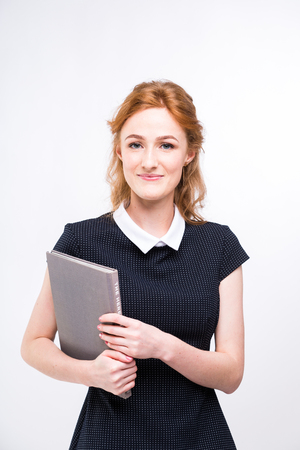 Beautiful girl with red hair and gray book in hands dressed in black dress on white isolated background.の写真素材