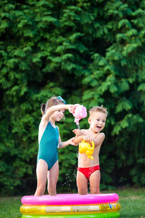 childhood summer games with water pool. Caucasian brother and sister play with plastic toys watering can pouring water splashing, inflatable round childrens bathroom. Summer hot holidays in swimsuits.の写真素材