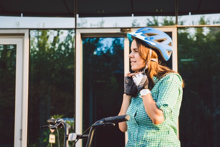 Theme to work on the bike. A young Caucasian woman arrived on environmentally friendly transport bike to the office. Girl in a bicycle parking office building in a helmet, gloves and shirt and jeans.の写真素材