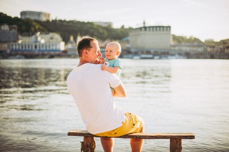 Subject recreation father and little son. Young caucasian dad sits on wooden bench overlooking the town Kiev and holding river of the Dnieperの写真素材