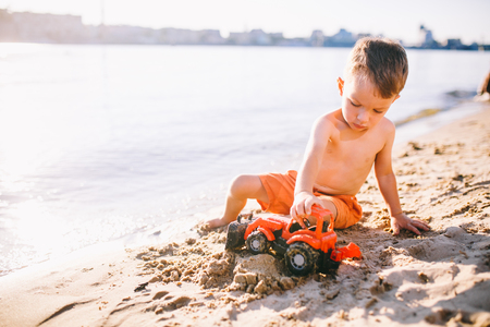 Subject construction and heavy industry. Abstraction child boy playing on the sand near the river in the summer toy red tractor model, excavator machine with a bucket.の写真素材