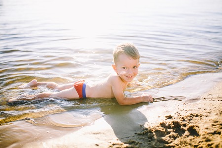 Theme is summer time and rest near the water. Little joyful Caucasian funny boy plays and enjoys in the river. The child is resting and swimming in the lake pond sandy beach.の写真素材