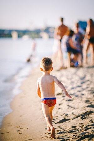 Theme summer outdoor activities near the river on the city beach in Kiev Ukraine. Little funny baby boy running along the river bank, jumping a splash of water sandy beach summer time.の写真素材