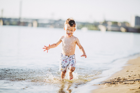 boy child playing makes splashes, beats hands on the water in the river at sunset of the dayの写真素材
