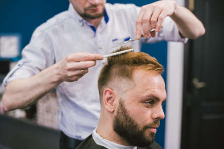 Barber Making Haircut Bearded Man In Barbershop. Professional stylist cutting client hair in salon. Barber using scissors and comb. Skillful hairdresser cutting male hair. Hair Care Service Concept.の写真素材